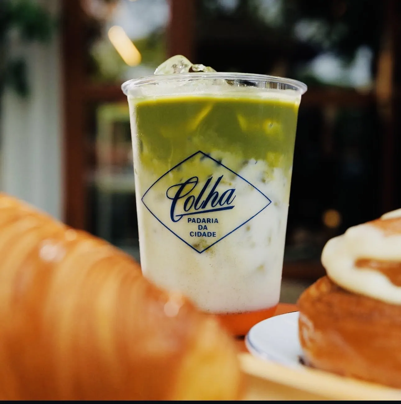 Matcha latte gelado com leite em copo da Colha, com croissant e cinnamon roll ao fundo